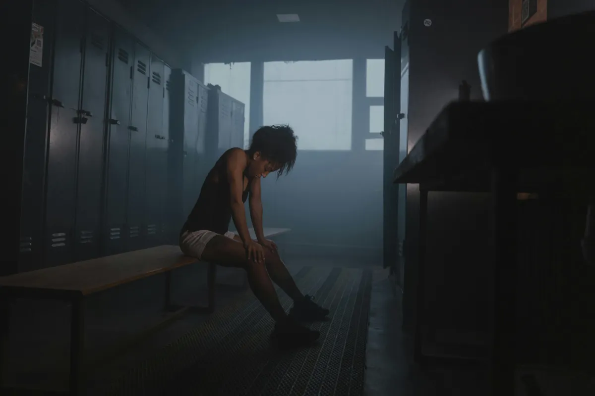 Young female athlete sitting alone on a locker room bench with head down, body slouched in fatigue, conveying emotional exhaustion and signs of sports burnout.