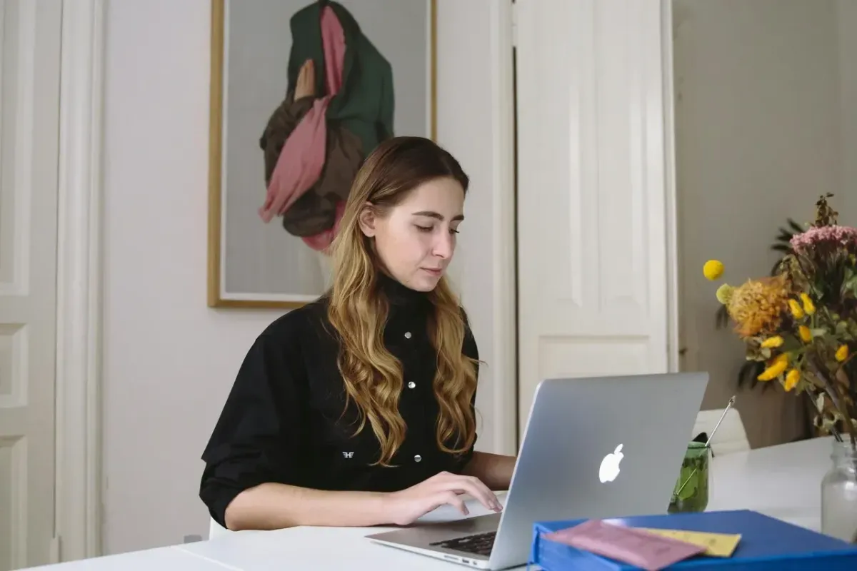 Woman working on a laptop at a white desk in a bright home office, wearing a black top, with artwork on the wall behind her and flowers on the side.