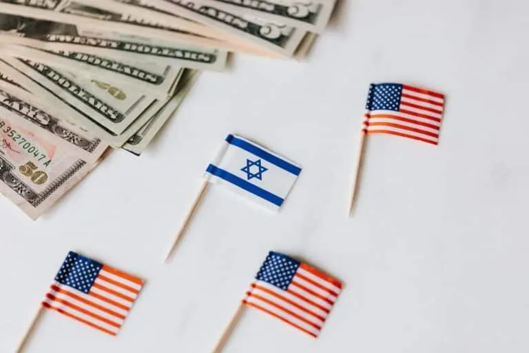 A stack of U.S. dollar bills is placed on the left side of the image. Surrounding it are five small flags on toothpicks: three U.S. flags and two Israeli flags, arranged on a white background. Explain flag symbolism Currency exchange rates