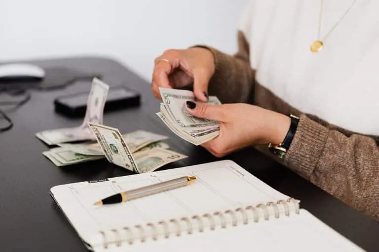 Person counting U.S. dollar bills at a desk with a planner and pen.