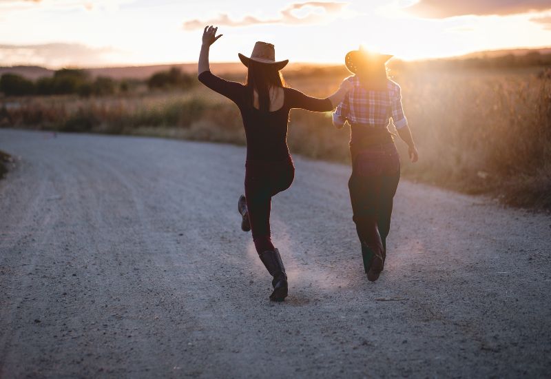 Two people wearing cowboy hats walking and playfully running down a rural dirt road at sunset.