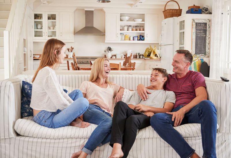 A family of four sits together on a light-colored, striped sofa in a bright, cozy living room that opens into a modern kitchen. From left to right: a teenage girl with long reddish hair, wearing a loose white sweater and blue jeans, sits cross-legged facing the others. Next to her, a blonde woman in a pale pink T-shirt and jeans leans back laughing warmly. Beside her, a young boy in a light gray T-shirt smiles broadly while leaning into a man who appears to be his father. The father, wearing a red striped T-shirt and jeans, has his arm around the boy and looks at him with a big smile.  The family appears relaxed and happy, sharing a lighthearted moment together. Behind them is a bright, tidy kitchen with white cabinets, open shelving with dishes and jars, a stainless steel range hood, and wooden dining chairs around a table. On the right side, a tall white refrigerator stands next to a chalkboard-style note board mounted on a cabinet door. The space is filled with natural light and soft neutral tones, creating a warm, comfortable home atmosphere.