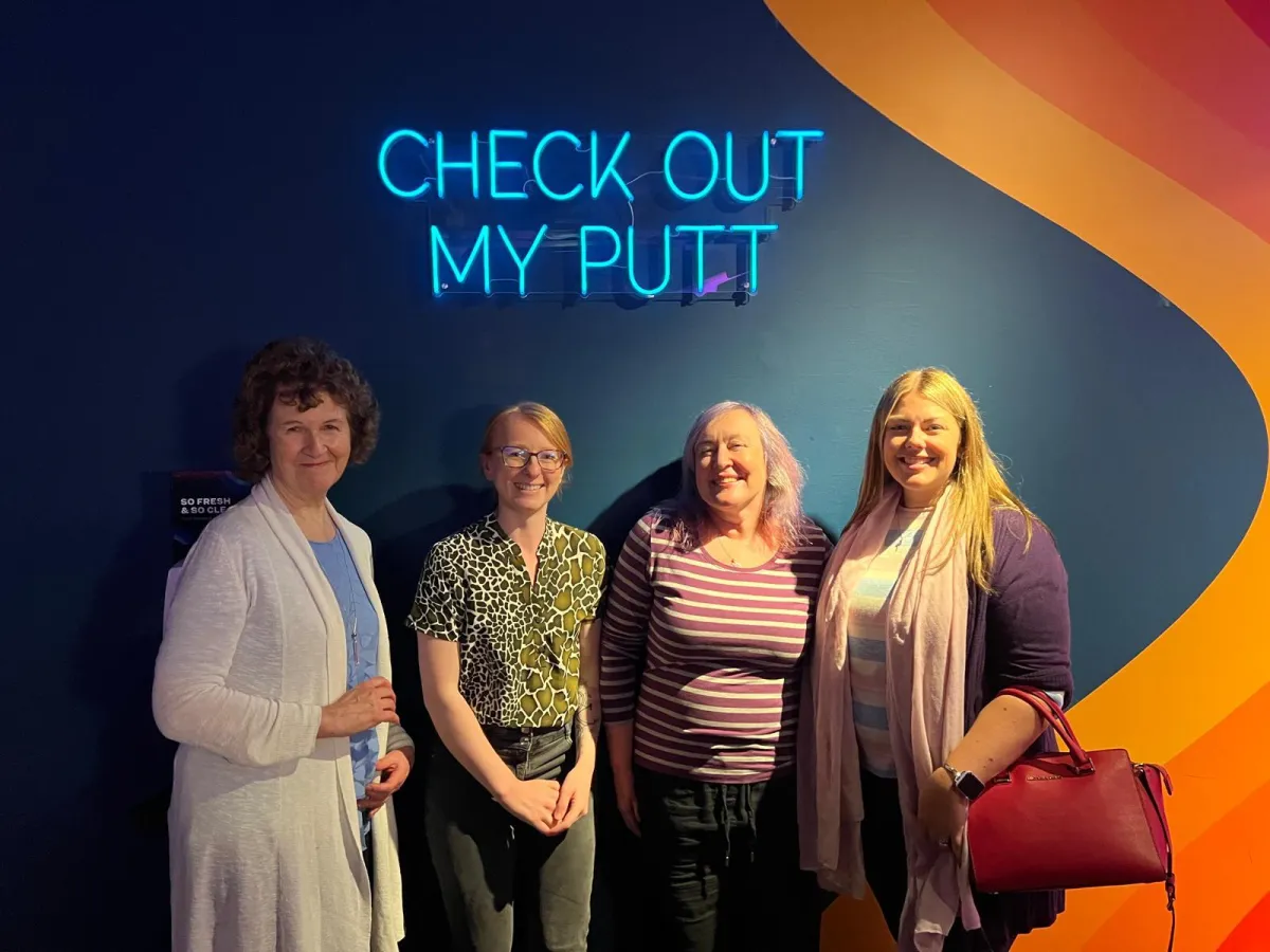 Four women posing in front of a neon sign that says “Check Out My Putt” at an indoor venue.