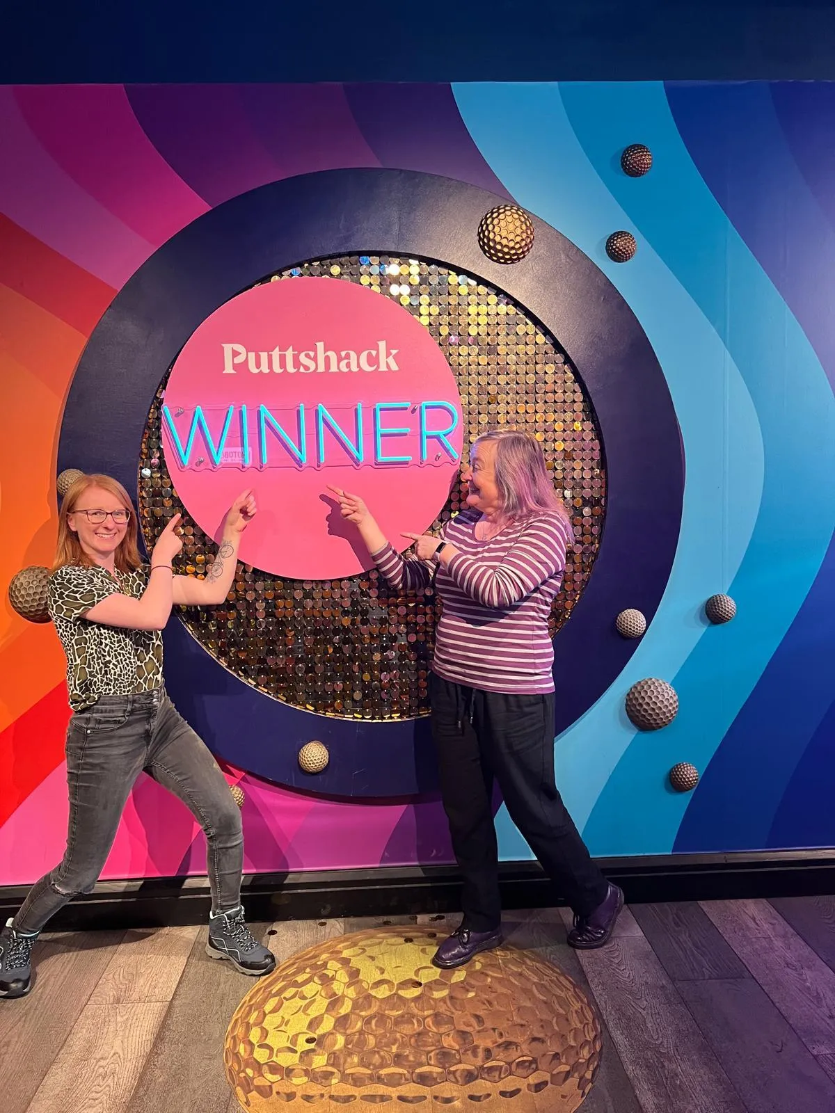 Two women celebrating in front of a colorful Puttshack Winner sign.