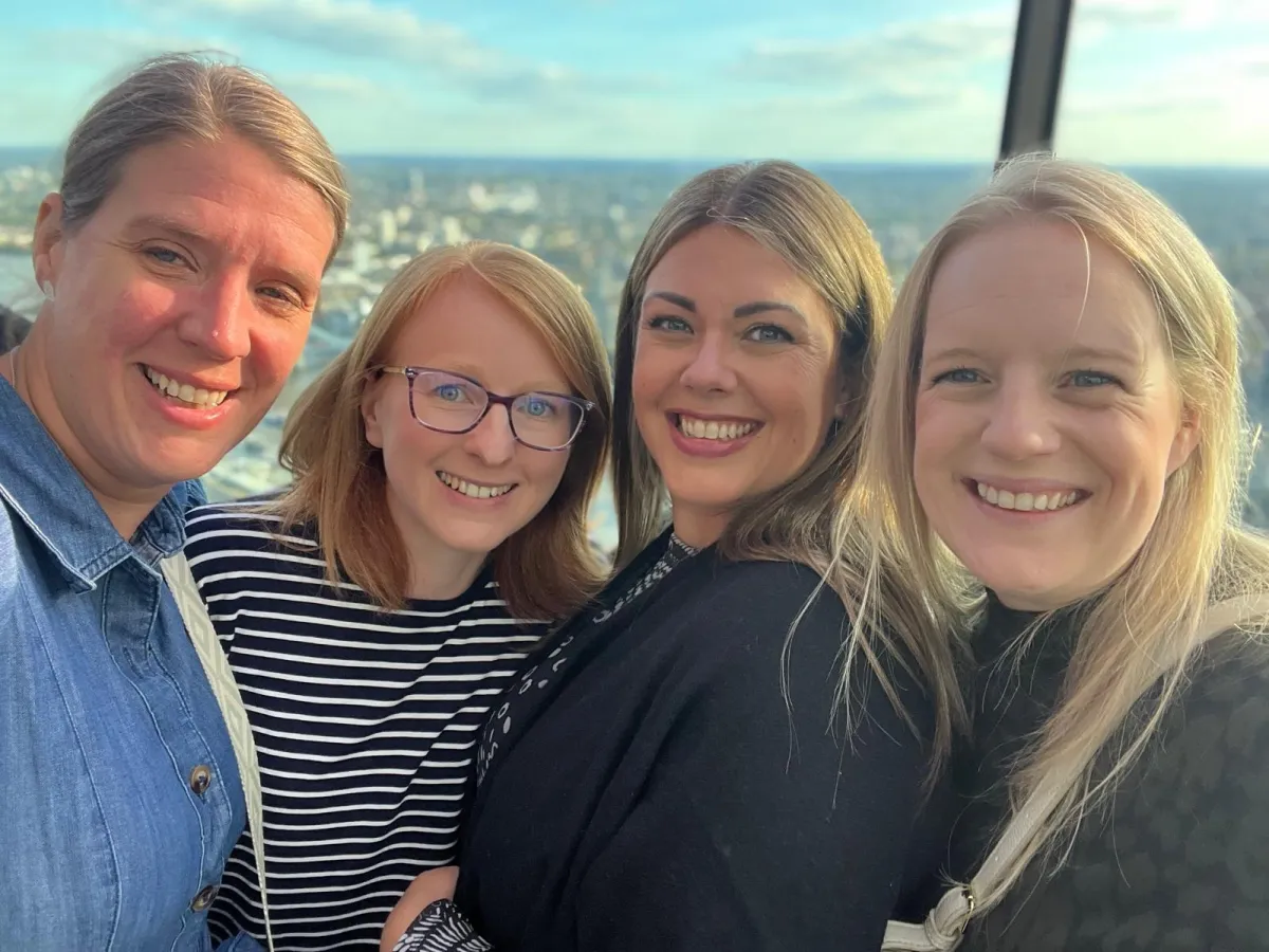 Close-up of four women smiling together outdoors with a sunny waterfront backdrop.