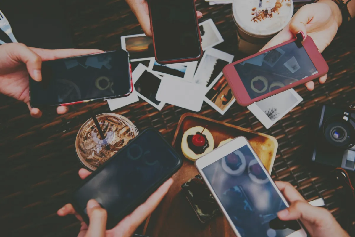 A bustling table scene from an overhead perspective where multiple hands are holding smartphones with black screens, possibly sharing content or capturing photos. Amidst the phones, the table is scattered with photographs, a camera, and refreshments including a frothy iced coffee and a dessert topped with a cherry. The atmosphere suggests a social gathering, likely focused on photography or digital sharing.