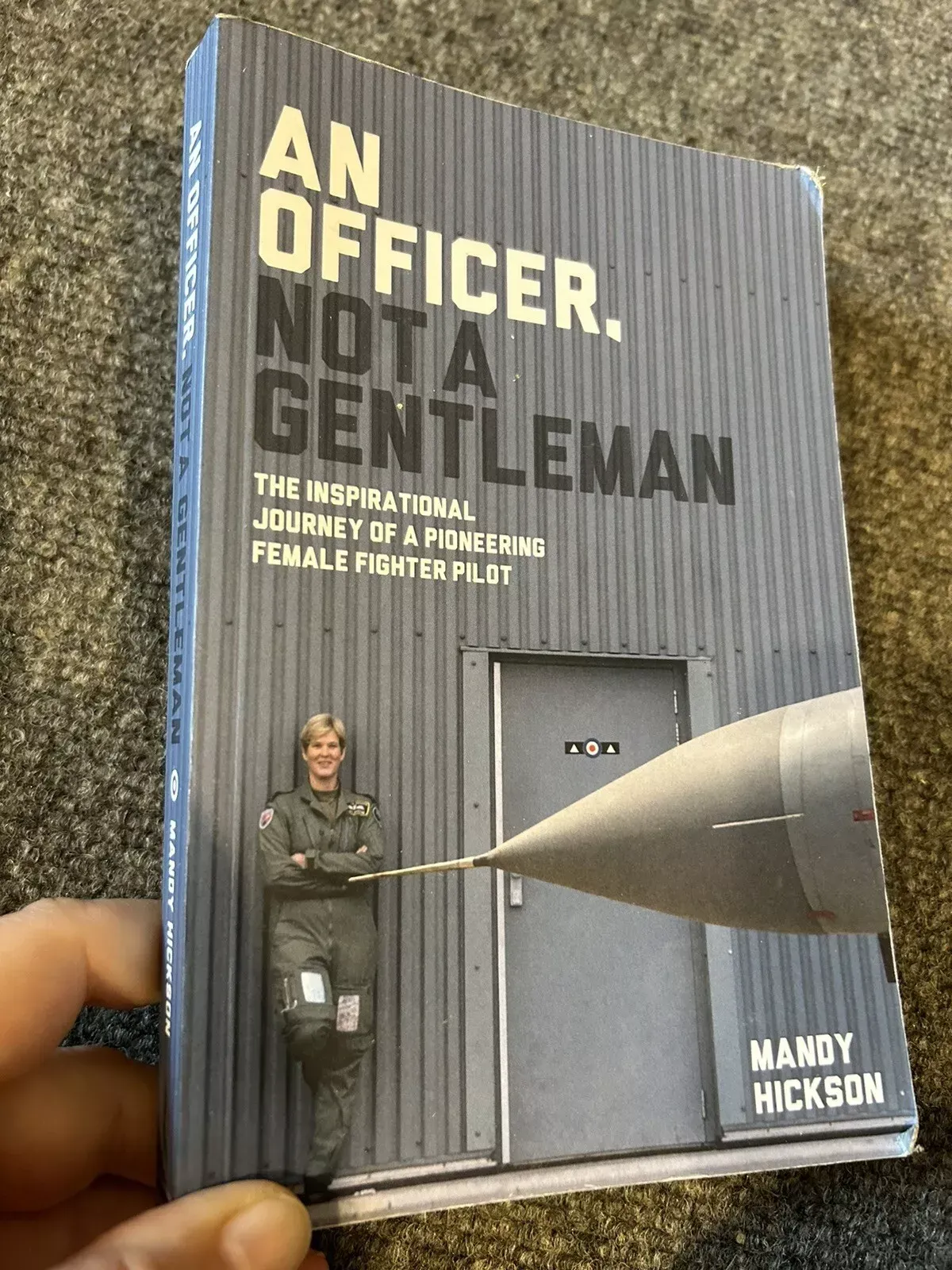 A paperback copy of the book An Officer, Not a Gentleman by Mandy Hickson is being held in someone's hand. The cover features a photograph of Mandy Hickson in her flight suit, leaning confidently against a military aircraft hangar, with the nose of a fast jet in the foreground. The book’s subtitle reads: The Inspirational Journey of a Pioneering Female Fighter Pilot.