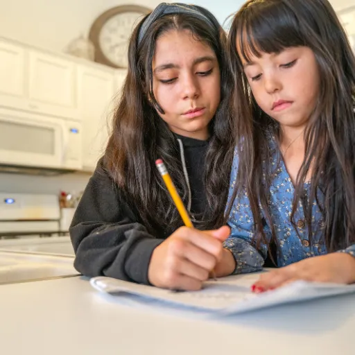 Older sister helping younger sister with homework at a kitchen table, showing support and care.