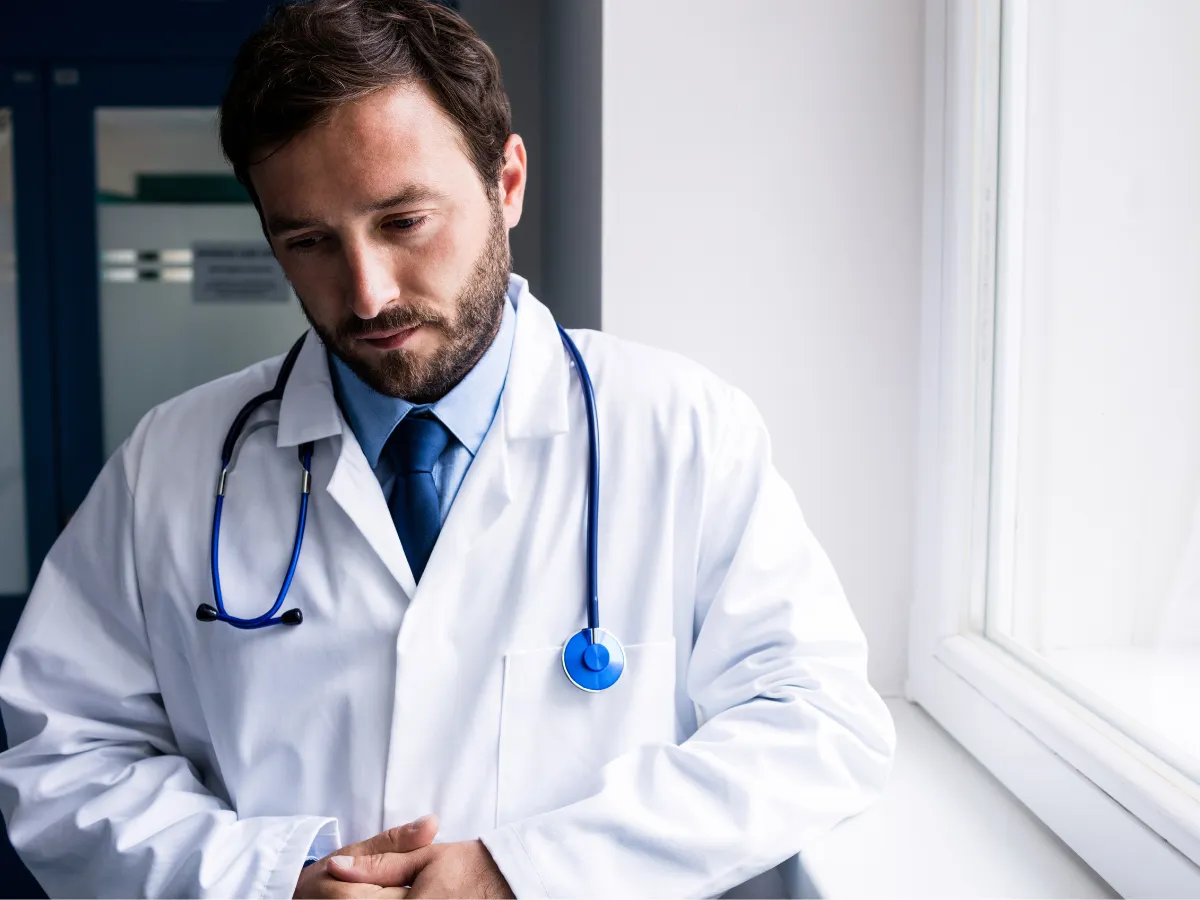 ad male physician in a white coat, sitting alone, reflecting; symbolic of burnout, emotional pain, and trauma in high-performing professionals.