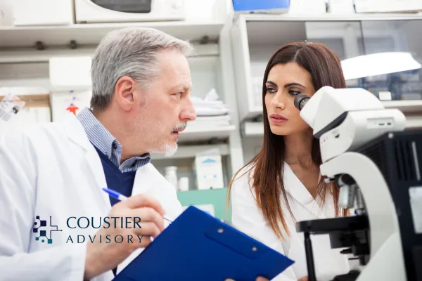 Two clinical researchers discuss trial data in a laboratory setting, with one reviewing notes on a clipboard and the other examining samples through a microscope.