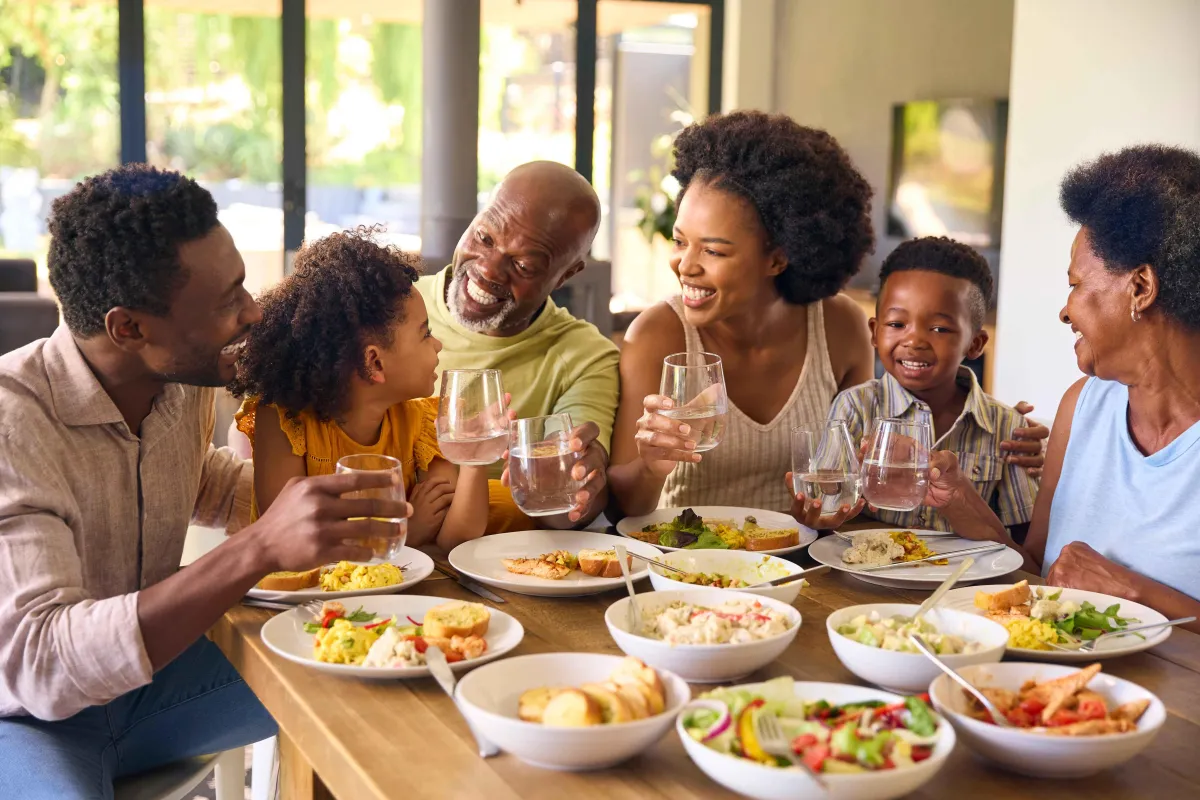 It shows a joyful, multi-generational family gathered around a table, enjoying a meal together. The positive, engaging atmosphere promotes the idea of family mealtime as a key moment for encouraging healthy eating habits.