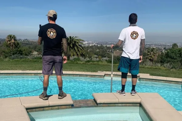 Two pool maintenance technicians standing at the edge of a backyard pool with tools in hand, overlooking a scenic city view and palm trees on a sunny day.