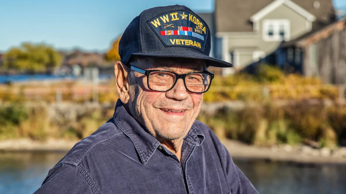 Smiling WWII and Korean War veteran wearing a blue veteran cap, sitting outdoors near water with houses in the background
