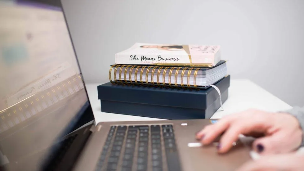 Hand typing on laptop with pile of business books in background