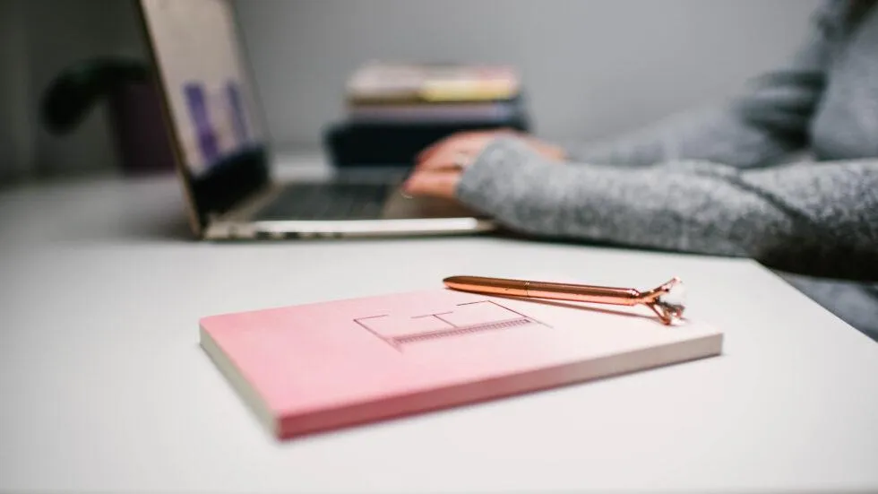 Notebook and pen with an "E" . Hands working on a laptop in the background