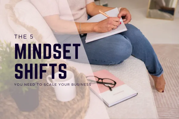 Woman writing in a notebook while sitting on a sofa, with a pink journal, black glasses, and coffee nearby. Overlay text reads “The 5 Mindset Shifts You Need to Scale Your Business”