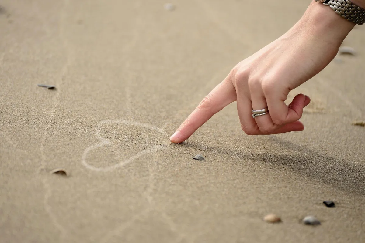 A close-up of a hand drawing a heart shape in the sand on a beach, symbolizing love, reflection, or peace.
