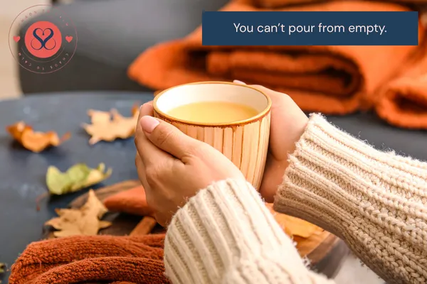 A parent’s hand gently holding a mug of tea, resting on a table in soft natural light, symbolising exhaustion, self-care, and the reminder that you can’t pour from an empty cup.