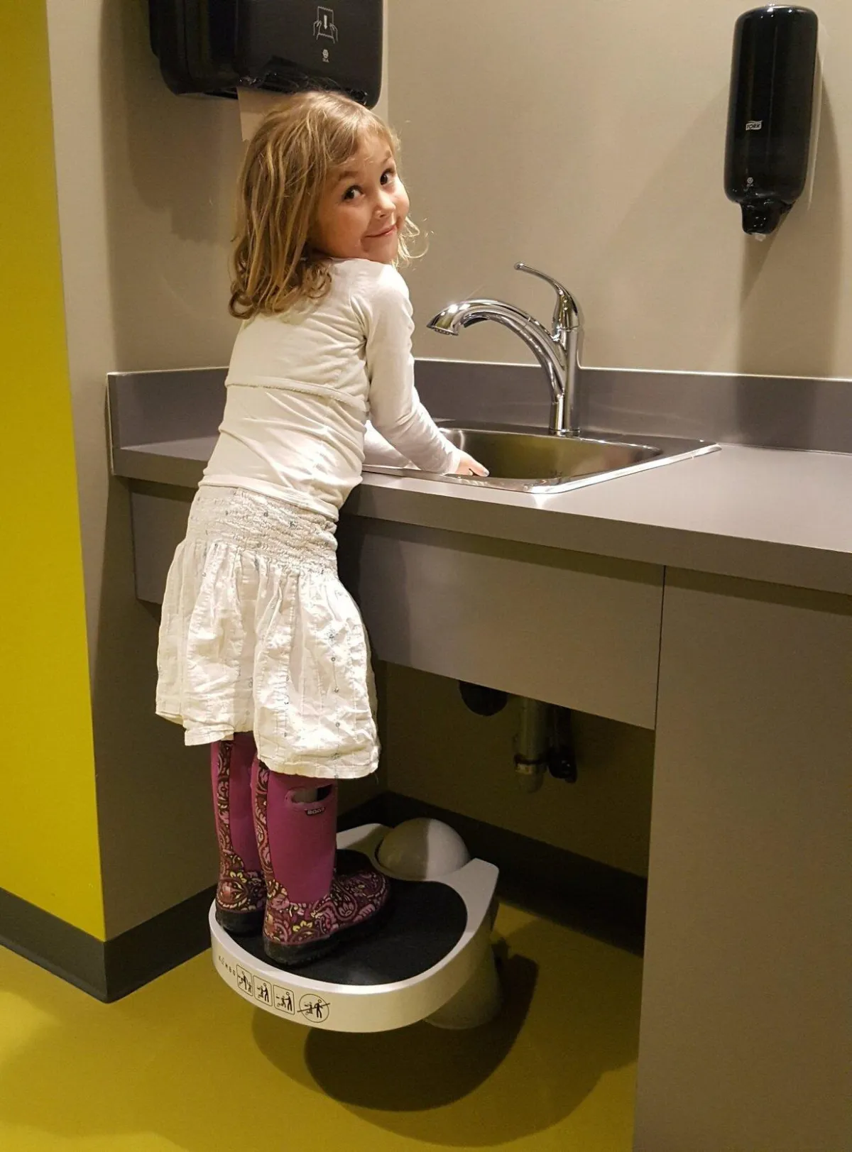 A toddler using a retractable step stool to elevate herself and wash her hands at a sink in a modern, clean bathroom, highlighting accessibility and child-friendly design