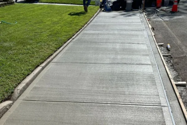 Fully renovated residential sidewalk with fresh broom-finished concrete and marked expansion joints, bordered by well-maintained grass and active work zone. Queens, New York