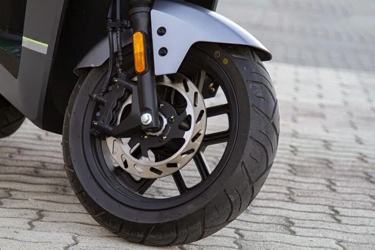 Sprednje kolo električnega skuterja VeloVoltage Titan s sodobnim zavorim sistemom CBS in vidno disk zavoro, parkirano na tlakovanem dvorišču.