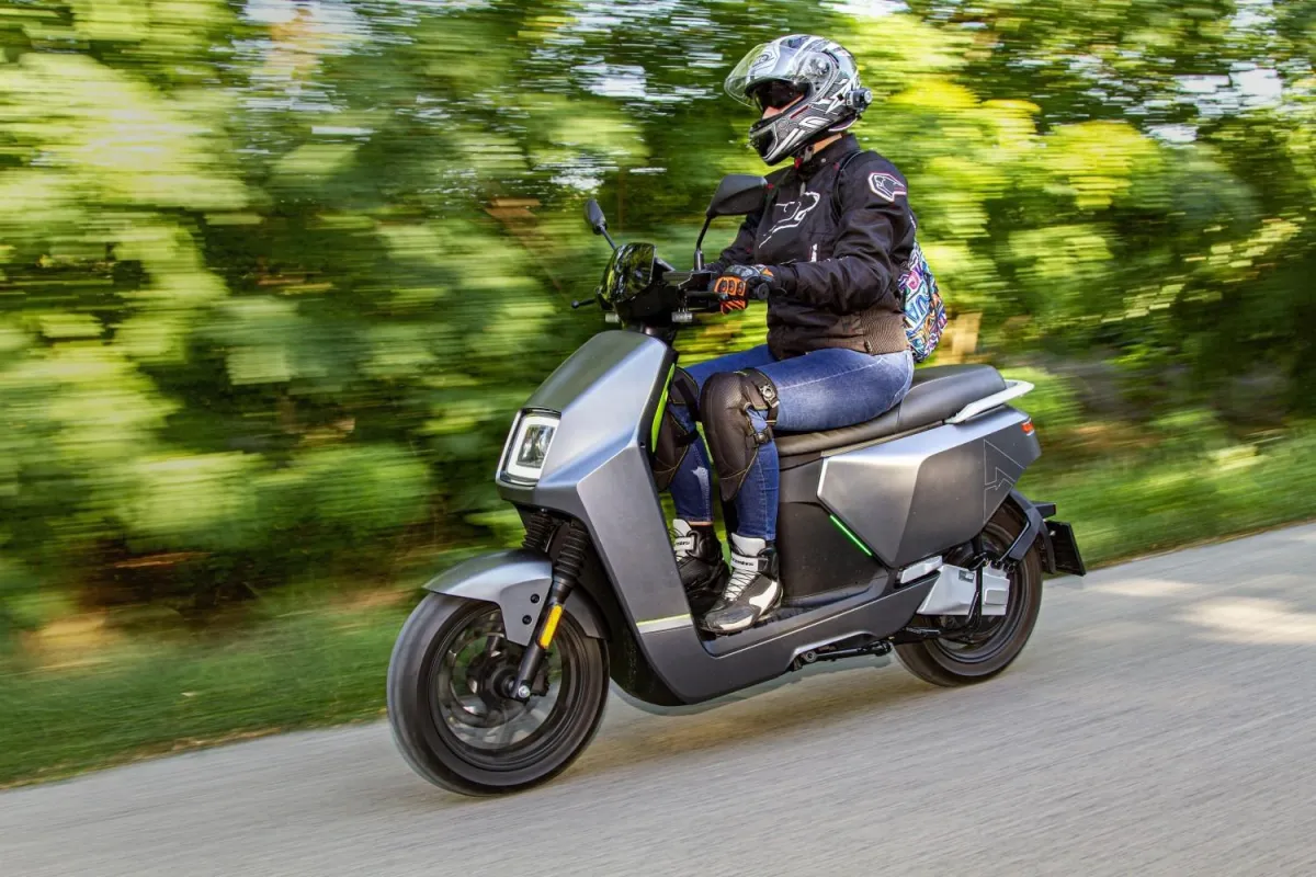 Voznik na električnem skuterju VeloVoltage Titan v temno sivi barvi med vožnjo po podeželski cesti, obdani z zelenjem, opremljen s čelado in motoristično jakno.