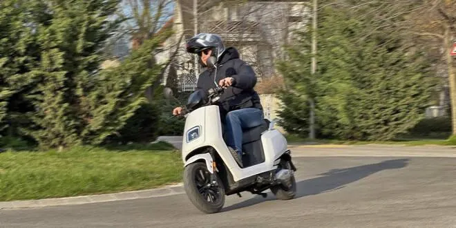 Oseba med vožnjo električnega skuterja VeloVoltage Prime v svetli barvi po mestni cesti, obkroženi z zelenjem in sončnim vremenom.