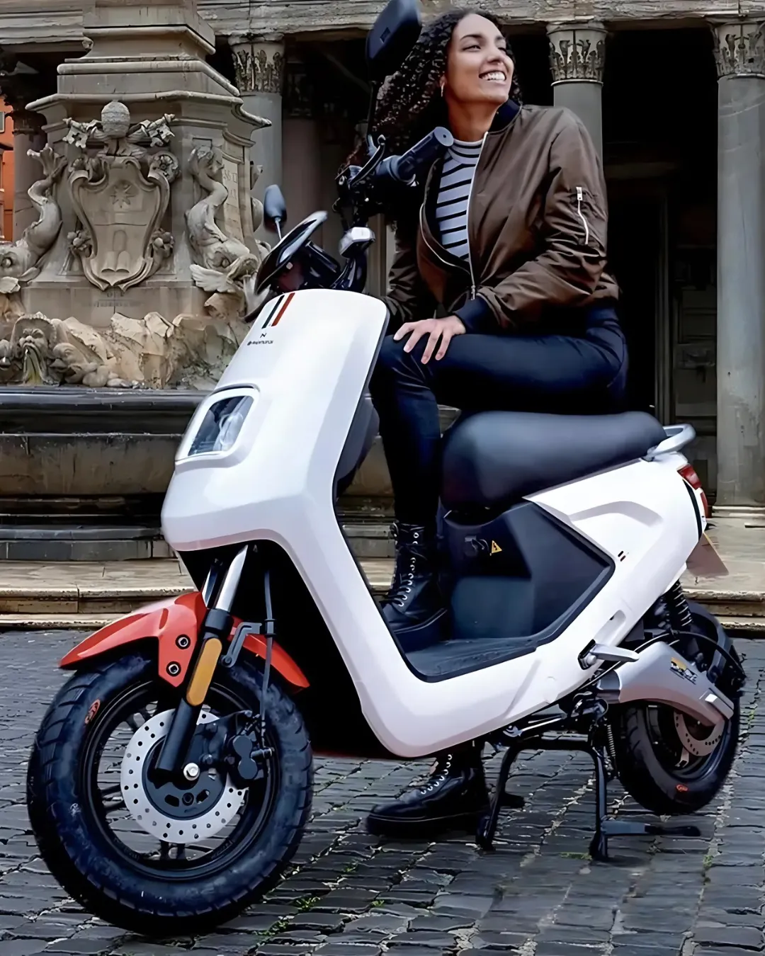 Nasmejana ženska sedi na belem električnem skuterju, parkiranem pred zgodovinsko zgradbo s kamnitimi okraski – popolna kombinacija sodobne mobilnosti in klasičnega mestnega ozadja.