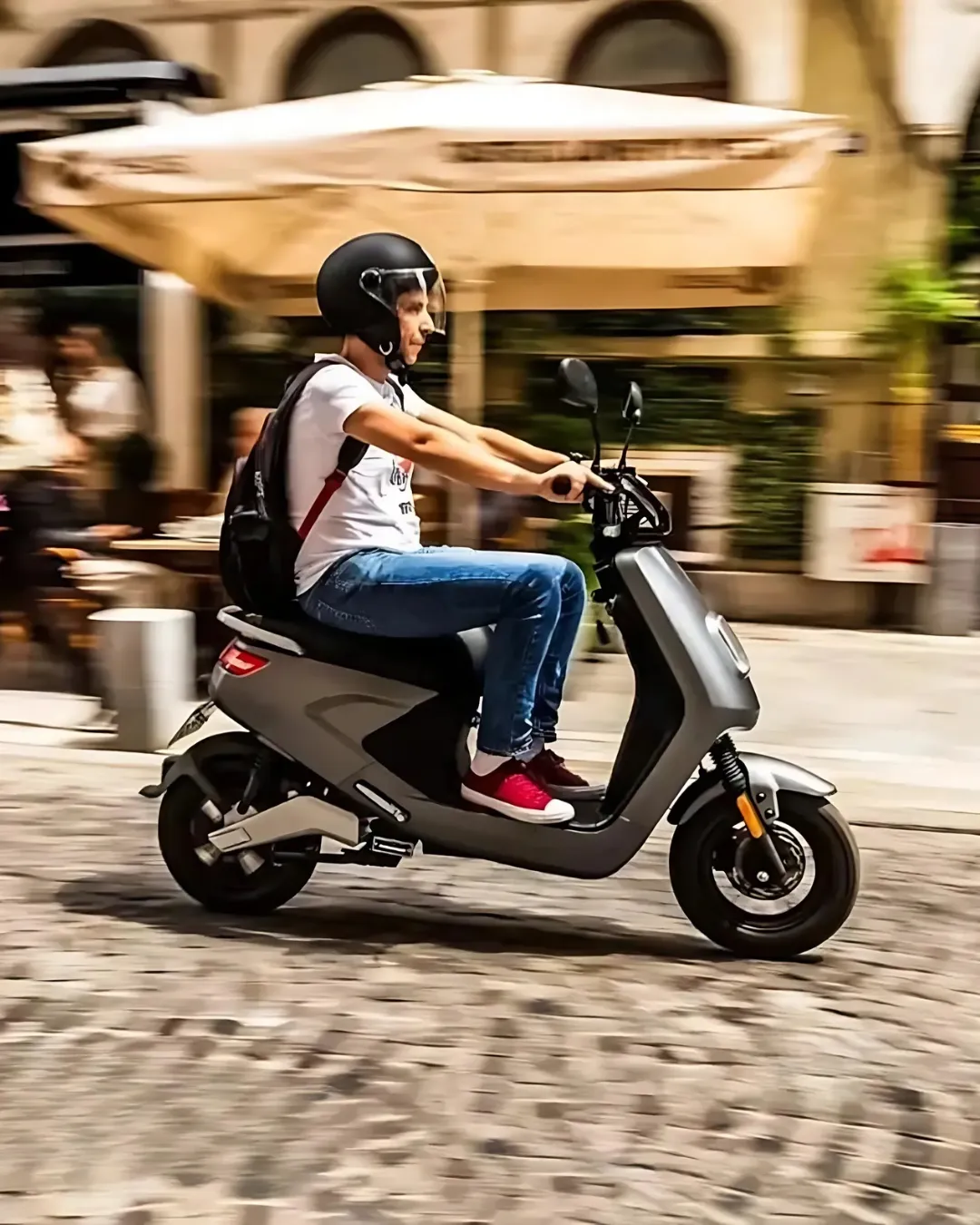 Moški z nahrbtnikom in čelado vozi temnosiv električni skuter po tlakovanih mestnih ulicah – dinamičen prizor urbane mobilnosti.