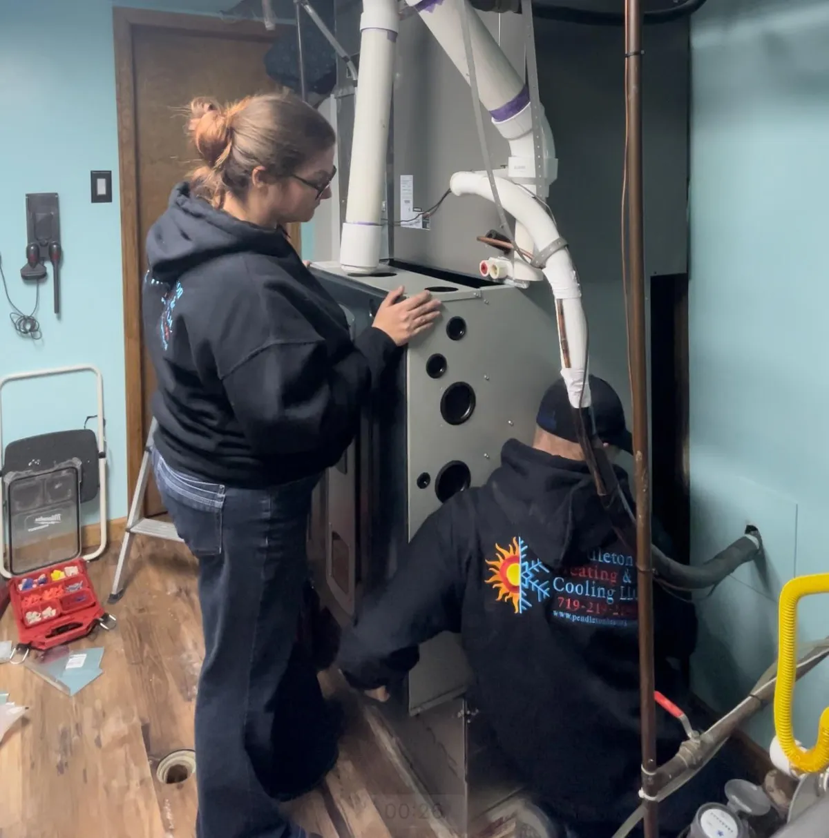 A woman and a man, both wearing black hoodies with the Pendleton Heating & Cooling LLC logo, are installing a furnace in a residential setting. The surrounding area has tools and equipment, indicating an HVAC installation in progress.