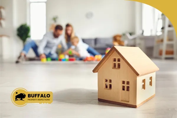 Wooden house model in the foreground on a living room floor, with a family playing together in the background of their new home.