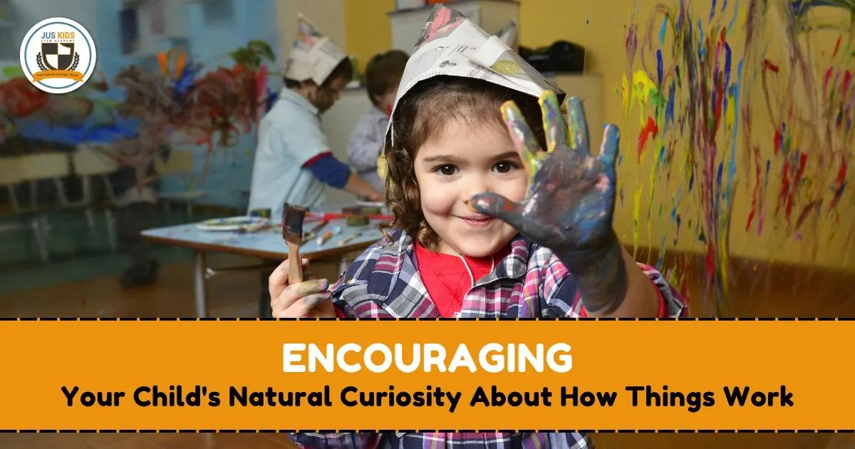 Smiling child wearing a paper hat shows a hand covered in colorful paint while holding a paintbrush, standing in front of a wall with creative artwork in a classroom. Other children are painting in the background. Text overlay reads ‘Encouraging Your Child's Natural Curiosity About How Things Work’ with the Jus Kids International School logo in the top left corner.