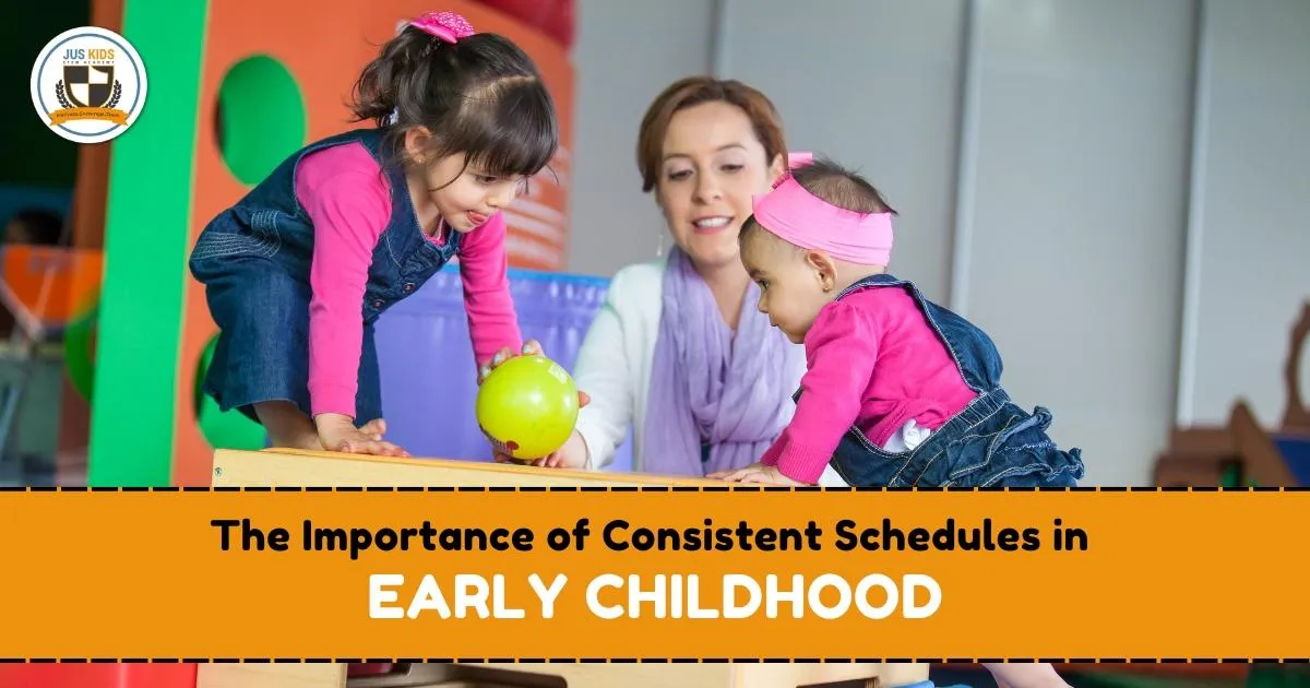 Two young children in matching pink shirts and denim overalls play together on playground equipment with a green ball, while a teacher supervises closely. Text overlay reads ‘The Importance of Consistent Schedules in Early Childhood’ and the Jus Kids International School logo appears in the top left corner.