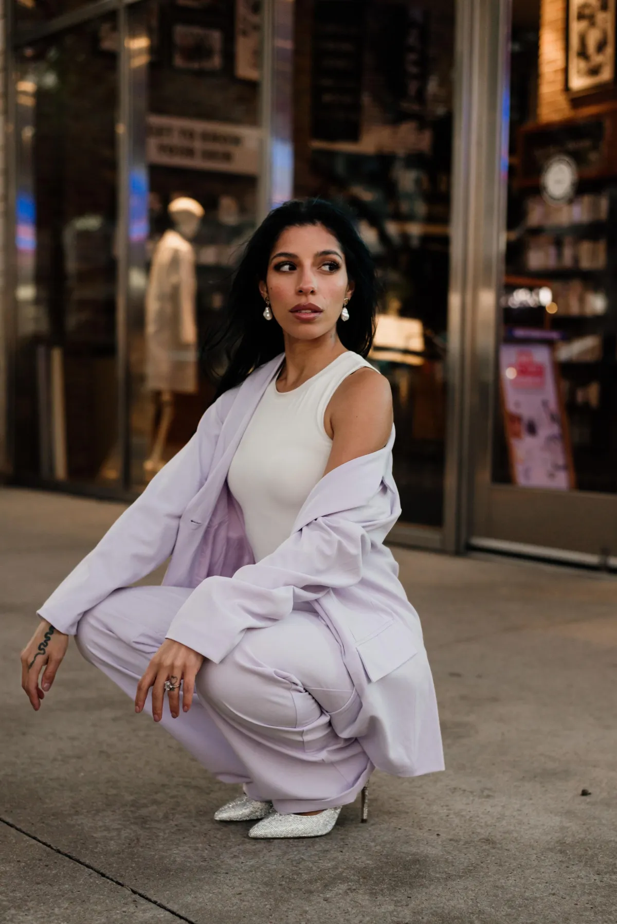 A confident woman in a stylish lavender suit and white top, crouching outdoors in front of a modern storefront, wearing pearl earrings and sparkling heels, exuding elegance and empowerment