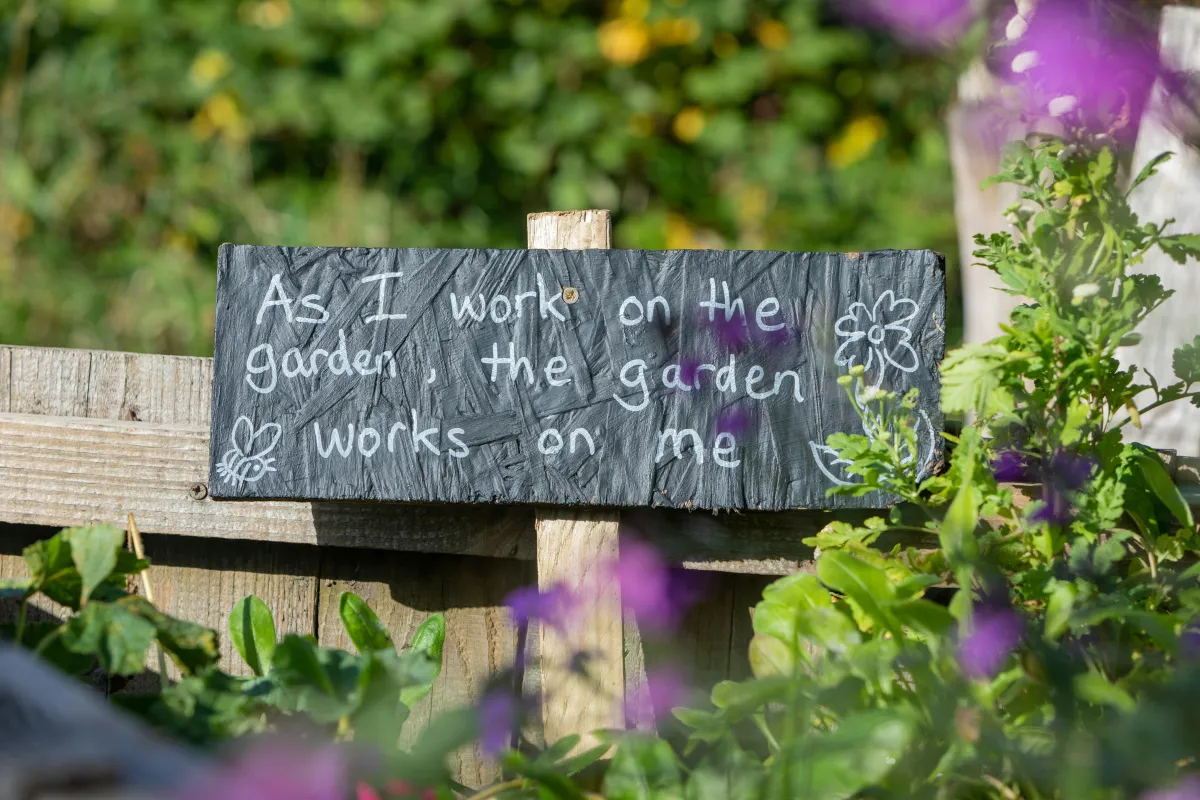 A small black chalkboard sign is placed inside a garden box.  The words, "As I work on the garden, the garden works on me" are written in white on the sign.