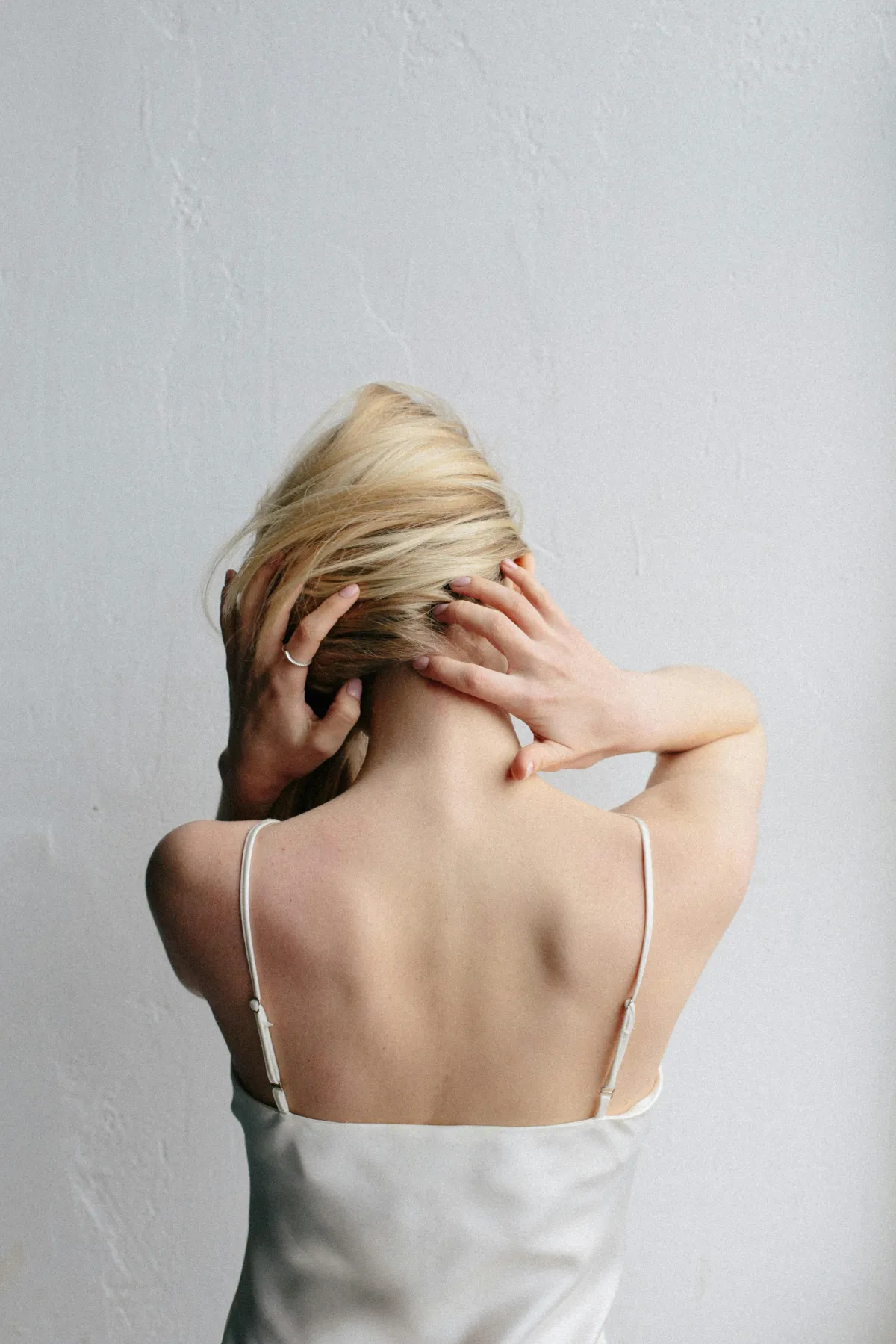 A woman is facing away from the camera, the back of her body in view.  She has blonde hair and has it pulled to the side as she holds onto her neck.  She's wearing a white tank top, and a white wall is in the background.