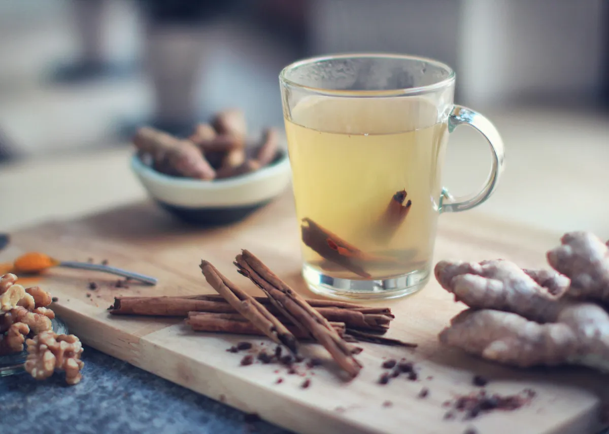 A glass teacup is on a table surrounded by large pieces of cinnamon and ginger