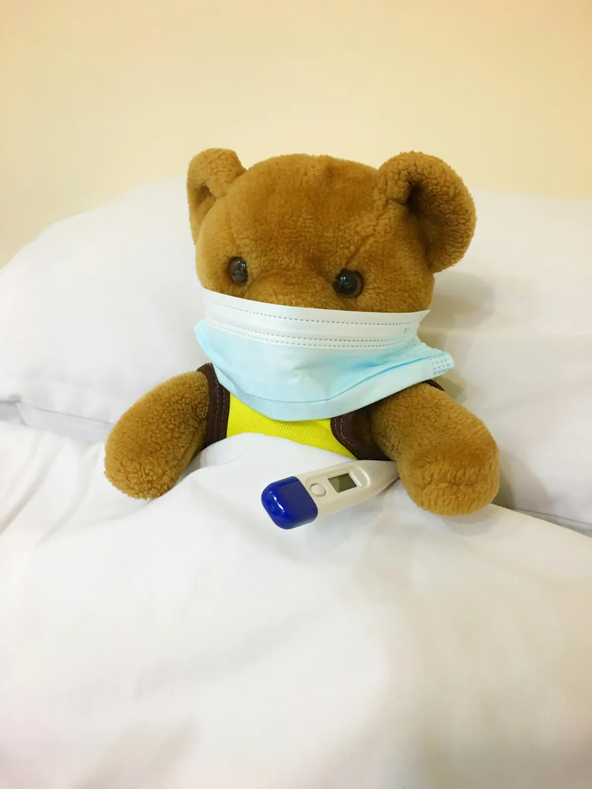 A teddy bear is on a bed, tucked under the covers.  It's wearing a blue surgical mask and is holding a small white thermometer.