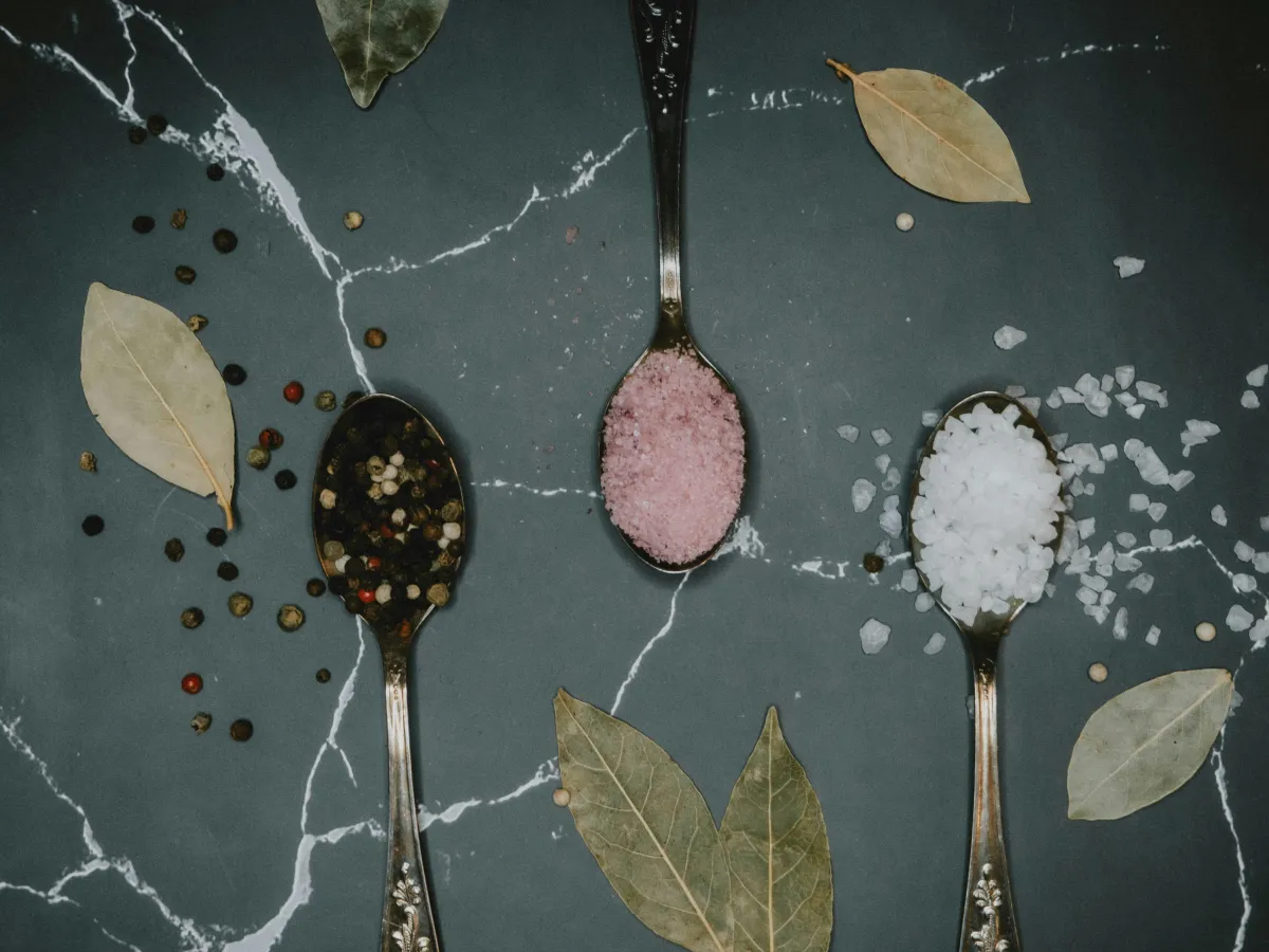 Three silver metal spoons lay atop a dark grey slab of granite.  One spoon is filled with black peppercorns.  One spoon is filled with pink rock salt, and the final spoon is filled with white rock salt.