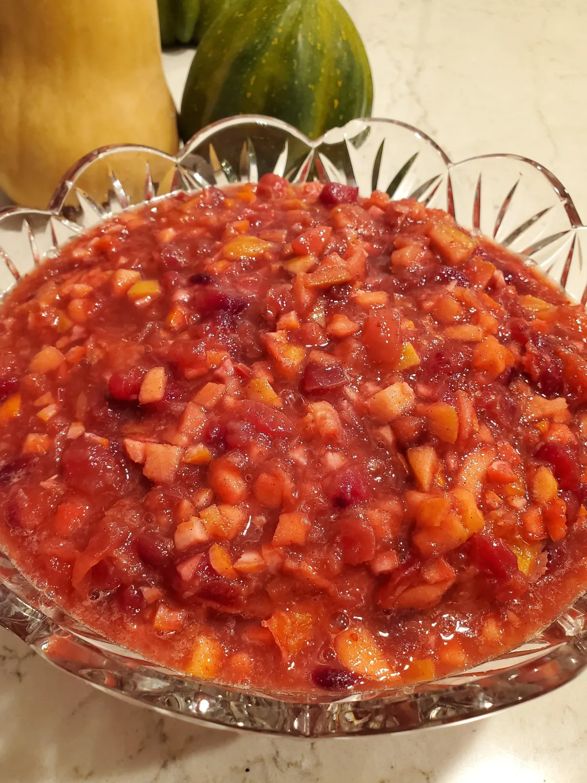 Fresh cranberry relish sits inside a crystal bowl, placed on a counter near squash
