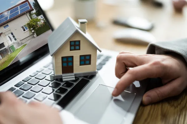 The image shows a person using a laptop to look at a house. The person is holding a small model house in their other hand. The laptop screen shows a picture of a house.