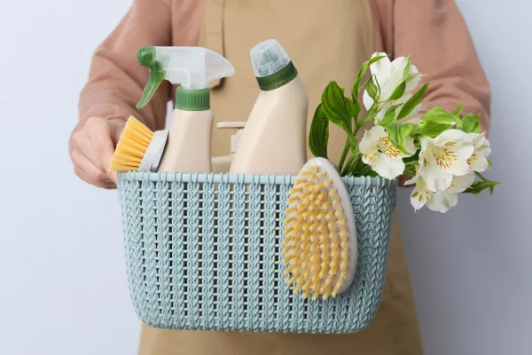 A person wearing an apron is holding a blue woven basket filled with cleaning supplies. The basket contains a spray bottle, a larger cleaning bottle with a green cap, a round brush, and a smaller yellow brush. There are also white flowers in the background.