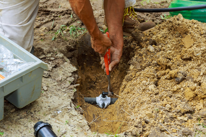 A worker kneels beside a dug-out trench and repairs an underground pipe joint, with loose soil and tools nearby.