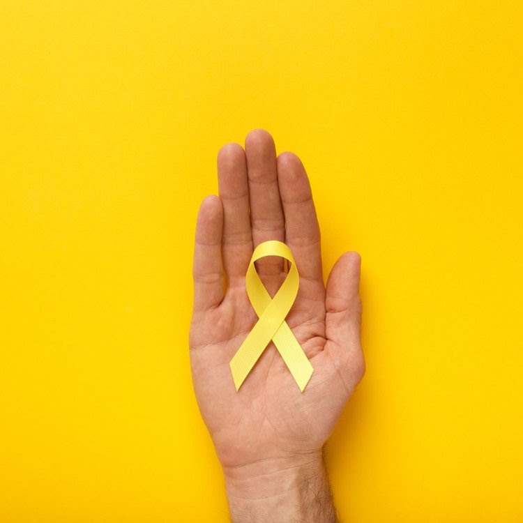 Mano abierta sosteniendo un lazo dorado sobre un fondo amarillo, símbolo mundial de la concientización sobre el cáncer infantil.