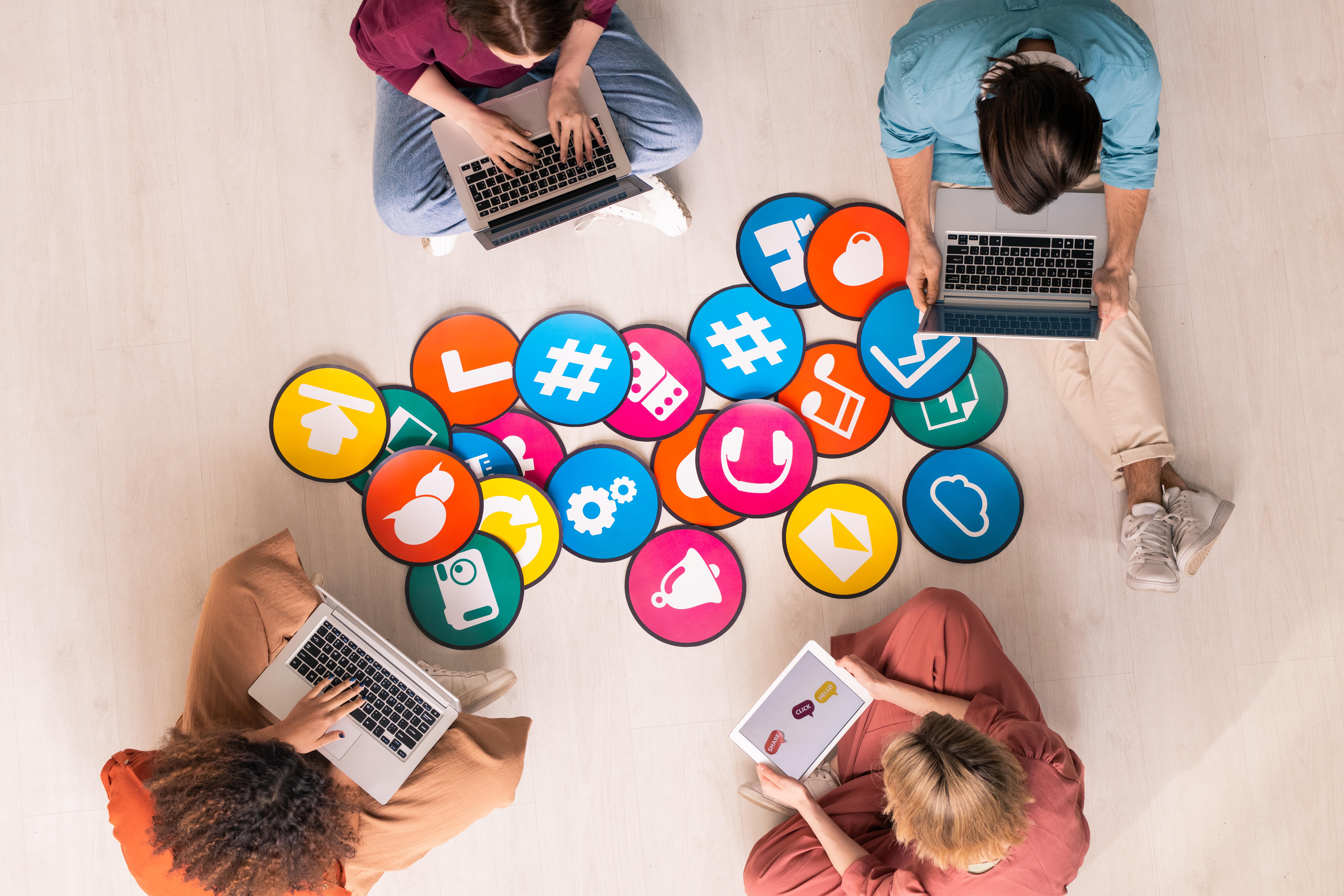 Overhead view of four people working on laptops and tablets, surrounded by colorful icons representing various social media platforms and digital marketing tools.