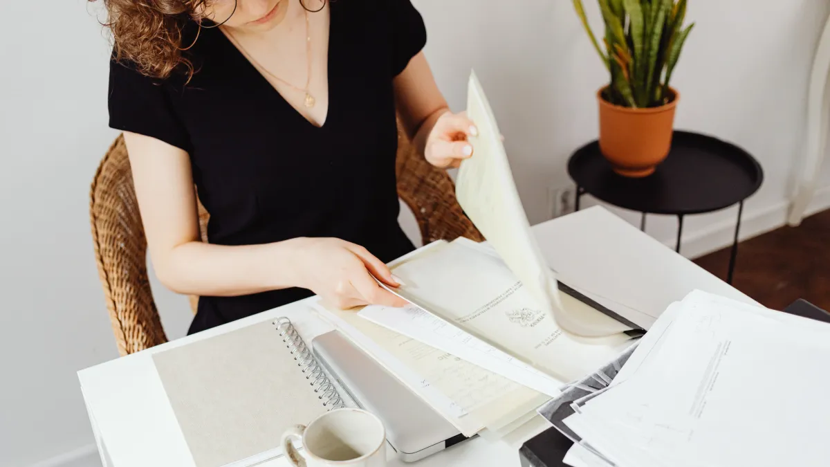 Woman in a black top sitting at a white desk, sorting through financial documents and papers in a bright home office, representing organizing essential financial records.