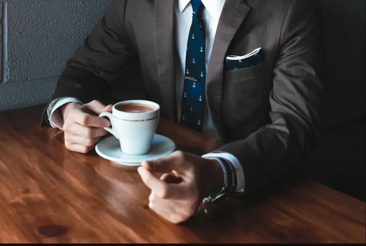 En affärsman i kostym som håller i en kopp kaffe vid ett träbord, symboliserar ett professionellt och personligt möte för att presentera offerter istället för att skicka dem.