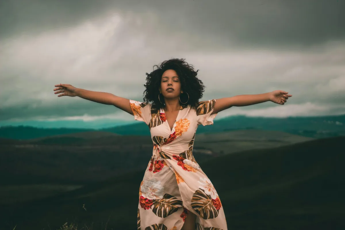 The image depicts a woman with curly hair standing in an open field with her arms outstretched and eyes closed, exuding a sense of freedom and tranquility. She is wearing a floral dress, and the background features rolling hills under a cloudy sky, creating a serene and natural atmosphere.