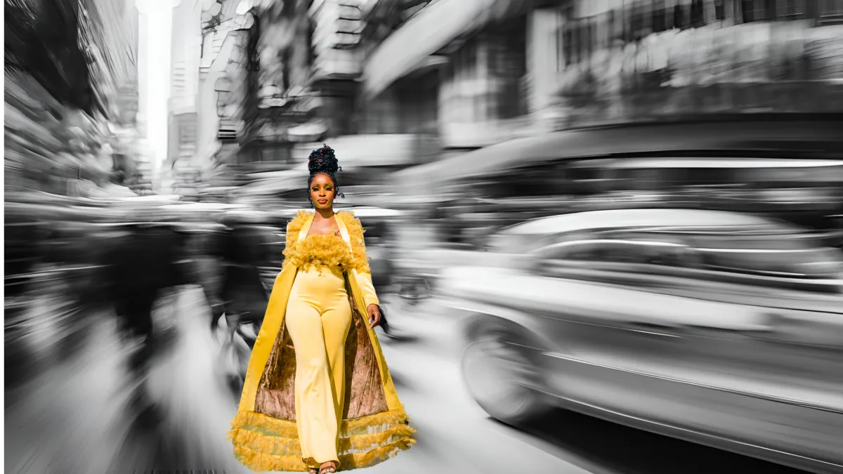 In the background a blurred busy city street with cars and people moving really fast and black and white conveying chaos. In the foreground, a beautiful black woman in full color to contrast the background, wearing a yellow pantsuit and long yellow cape, walking confidently and calmly through the chaos directly toward the viewer.