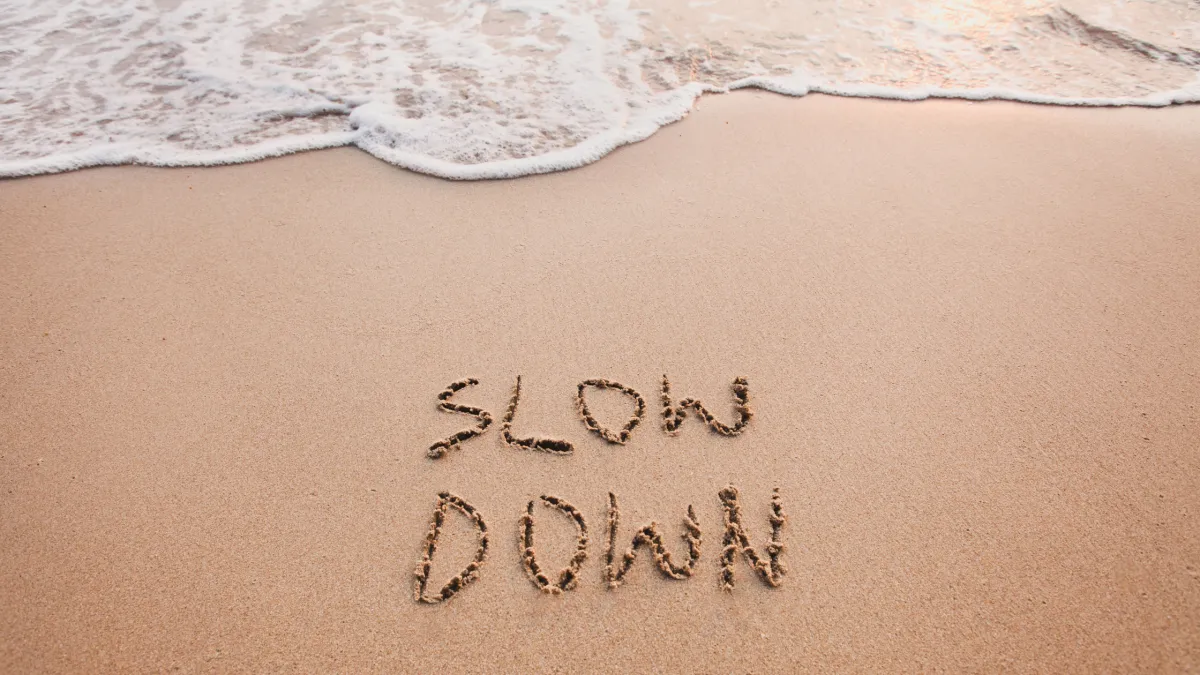 A sandy beach with a gentle wave coming in from the top of the image. The words “slow down” carved in the sand in the center of the bottom of the image.
